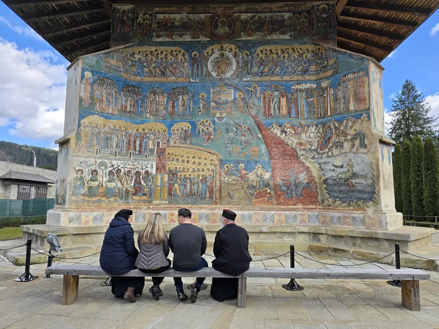Four people on a bench looking up at the painted west wall of Voroneț Monastery